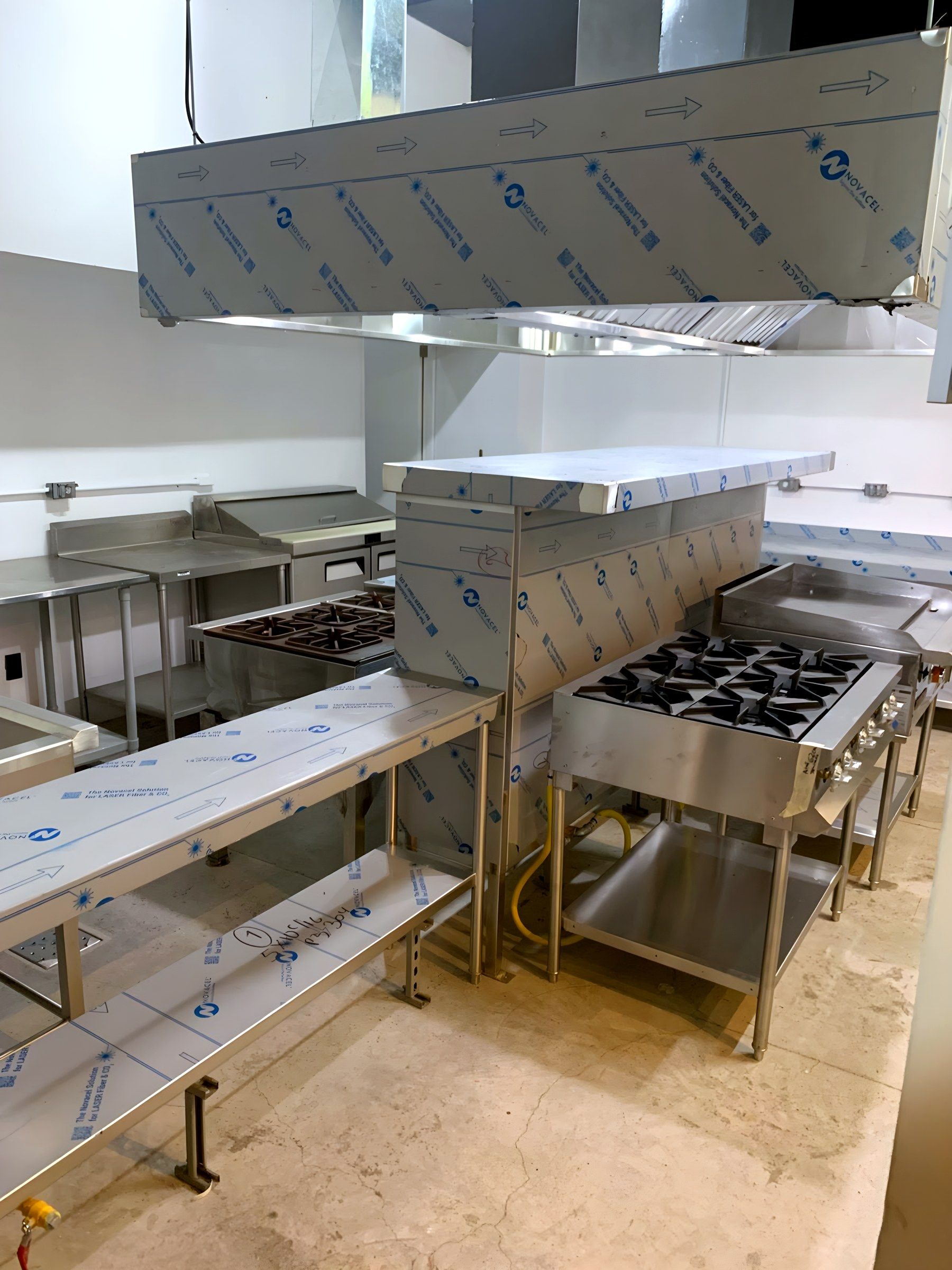 Cocina abierta con campana central, mesas de trabajo y estufas, aún con película protectora.