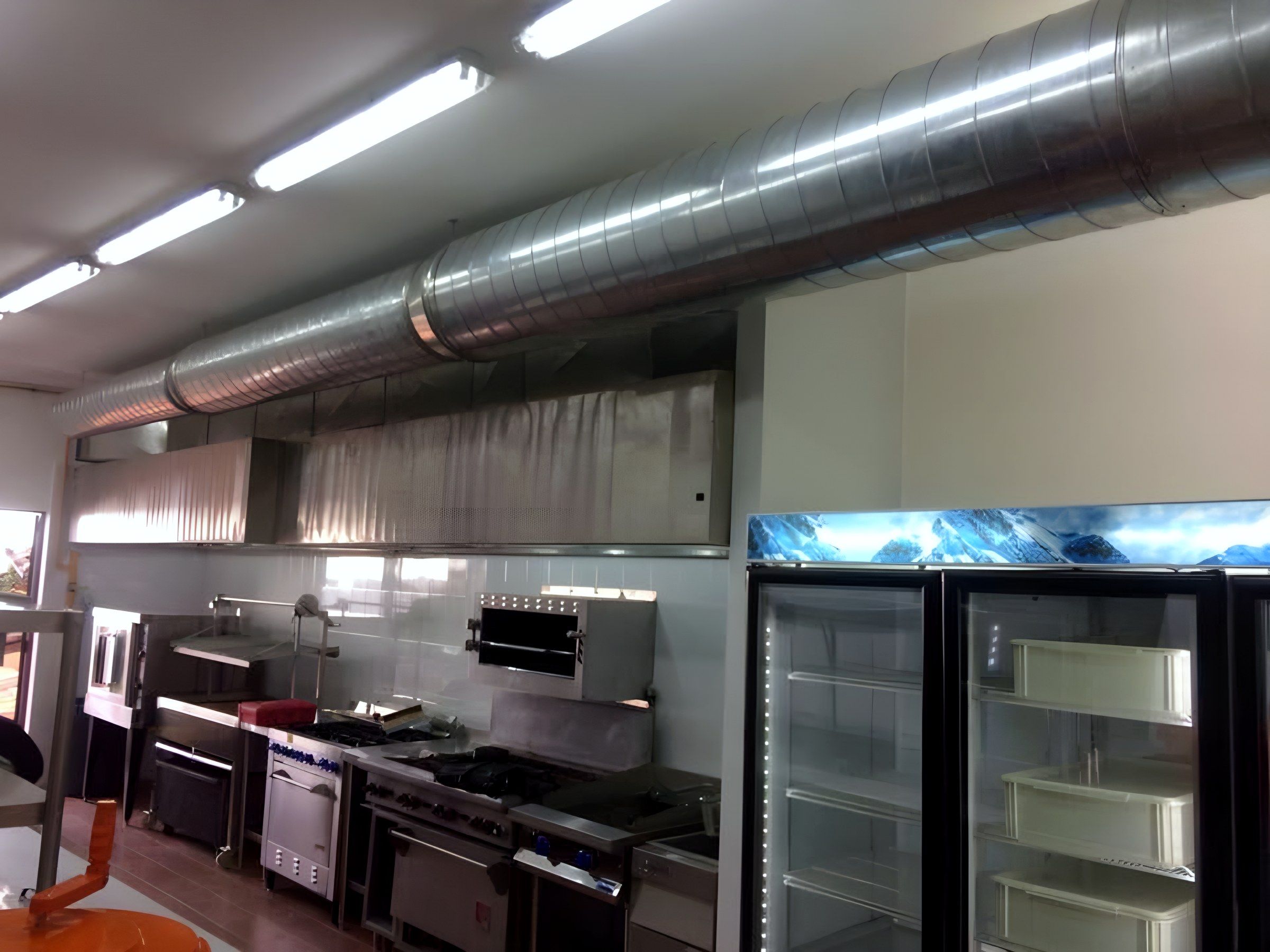 Línea de cocción bajo campana con ducto aparente, junto a refrigeradores de puertas de cristal.